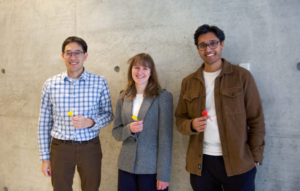Three people stand in front of a concrete wall. Timothy Chan stands on the left wearing a shirt with a blue windowpane pattern and is holding a yellow dart, Rachael Walker stands in the centre wearing a grey blazer and is holding a yellow dart and Craig Fernandes stands on the right wearing a brown coat and is holding a red dart.